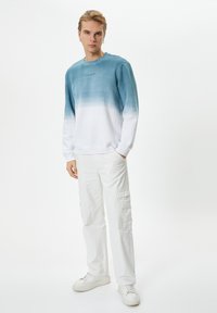 Felpa leggera con una sfumatura che va dal verde acqua al bianco, con collo a giro e un branding discreto sul petto, abbinata a pantaloni cargo bianchi.
