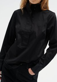 Schwarze Hemdbluse mit hohem Kragen, aus einem glatten Stoff gefertigt, mit verdeckter Knopfleiste und langen Ärmeln. Minimalistisches Design.