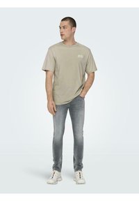 Tricou bej cu mânecă scurtă, cu un logo imprimat, asortat cu blugi slabuți gri deschis și adidași albi. Material neted și design casual.