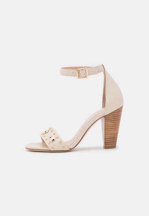 Sandale à talons hauts en cuir beige avec une large bride au niveau des orteils ornée de clous dorés, une bride de cheville avec une boucle et un talon en bois.