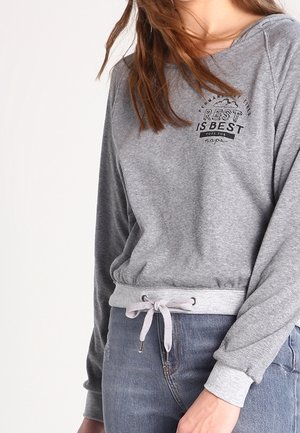 Femme portant un sweat-shirt gris à manches longues avec le texte "Rest is Best" et un cordon de serrage à la taille, associée à un jean bleu.