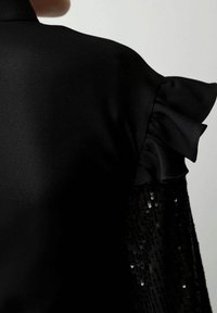 Schwarzes Kleid mit satinartigem Oberteil, Rüschenärmeln und einer mit Pailletten besetzten Stofflage, die Textur und Schichtung in dunklen Tönen betont.