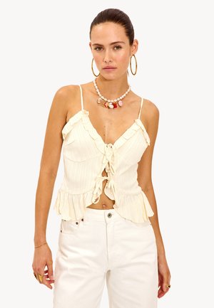 Femme portant un débardeur crémeux à volants avec un nœud sur le devant, un pantalon blanc, de grandes boucles d'oreilles en créoles, un collier en perles avec des charms colorés et des bagues en or.