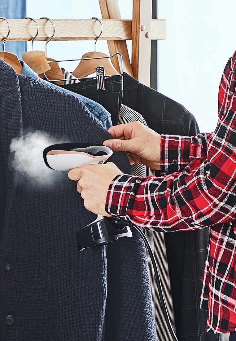 Draagbare stofstomer, zwart en wit, in gebruik op een donker gemêleerd cardigan. Stoom komt vrij, met kledingrekken en hemden op de achtergrond.