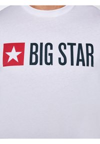 Fehér póló, melyen egy piros négyzet látható egy fehér csillaggal, utána vastag fekete betűkkel a "BIG STAR" felirat a mellkas közepén.