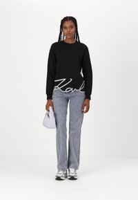 Felpa nera con un prominente logo bianco "Karl" in basso, abbinata a jeans dritti blu chiaro e una piccola borsa bianca.