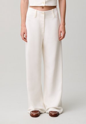 Donna che indossa pantaloni bianchi larghi a vita alta e mocassini marroni, in piedi davanti a uno sfondo chiaro e uniforme.