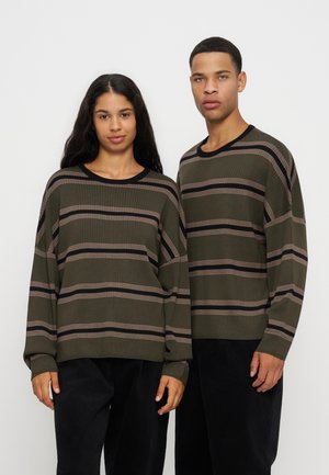 Un homme et une femme se tiennent côte à côte portant des pulls vert olive assortis avec des rayures horizontales noires et beiges et des pantalons noirs.