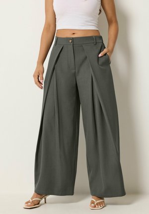 Donna che indossa pantaloni grigi a vita alta, a gamba larga con pieghe, un crop top bianco e sandali bianchi con il tacco, in piedi con una mano in tasca.
