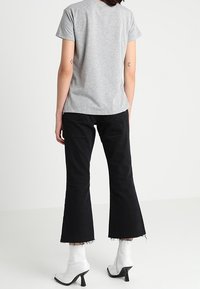 Person iført en grå kortærmet skjorte, sorte cropped flare jeans med rå kanter og hvide stilettokampagne ankelstøvler, står op ad en hvid baggrund.