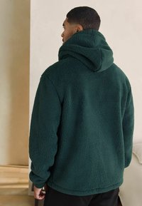 Ciemnozielona polarowa bluza z kapturem o teksturowanej, miękkiej powierzchni. Posiada wysoki kołnierz i przymocowany kaptur, z luźnym, oversize'owym krojem.