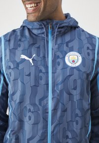 Giacca blu con cappuccio e motivo numerico; presenta un logo Puma e il badge del Manchester City; texture liscia e chiusura con cerniera.