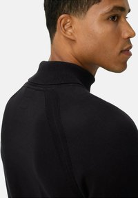 Junger Mann in einem schwarzen, gerippten Rollkragenpullover, der zur Seite schaut und die Profil- sowie Rückendetails des Kleidungsstücks zeigt.