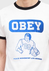 Άσπρο t-shirt με μπλε κείμενο "OBEY,", εικονογράφηση ποδοσφαιριστή που κρατά μια υδρόγειο, και κείμενο "PEACE MOVEMENT LOS ANGELES" από κάτω.
