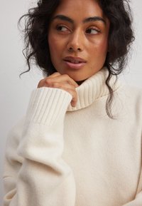 Krems krāsas augstais kakls džemperis, kas izgatavots no mīksta adījuma auduma. Iezīmējas ar ribotām aprocēm un kaklu; brīva piegriezuma, maiga tekstūra un smalka spīduma.