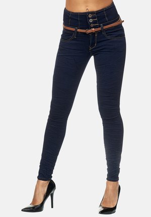 Vrouw die donkerblauwe skinny jeans met een hoge taille draagt, een bruine riem en zwarte hoge hakken, staand met licht gespreide benen.