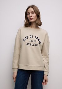 Sweat-shirt beige à col rond, avec un texte bleu foncé : "RUE DE PARIS No.9 ATELIER." Tissu doux, coupe décontractée et poignets côtelés.