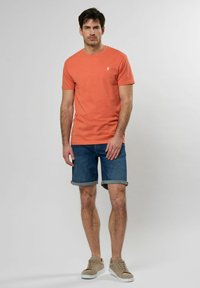 Oranžové bavlněné tričko s kulatým výstřihem, spárované s džínsovými šortkami s ohrnutými okraji. Modelka má na sobě béžové tenisky.