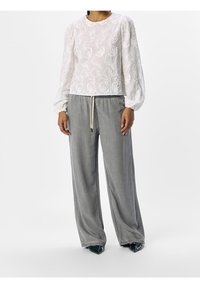 Blouse blanche brodée avec manches longues bouffantes associée à un pantalon ample gris clair et des chaussures noires à bouts pointus. Palette de couleurs neutres.