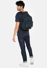 Mochila de tela azul con la parte superior redondeada, bolsillo frontal y base marrón; el modelo lleva una camiseta azul marino y jeans oscuros. Zapatillas blancas visibles.