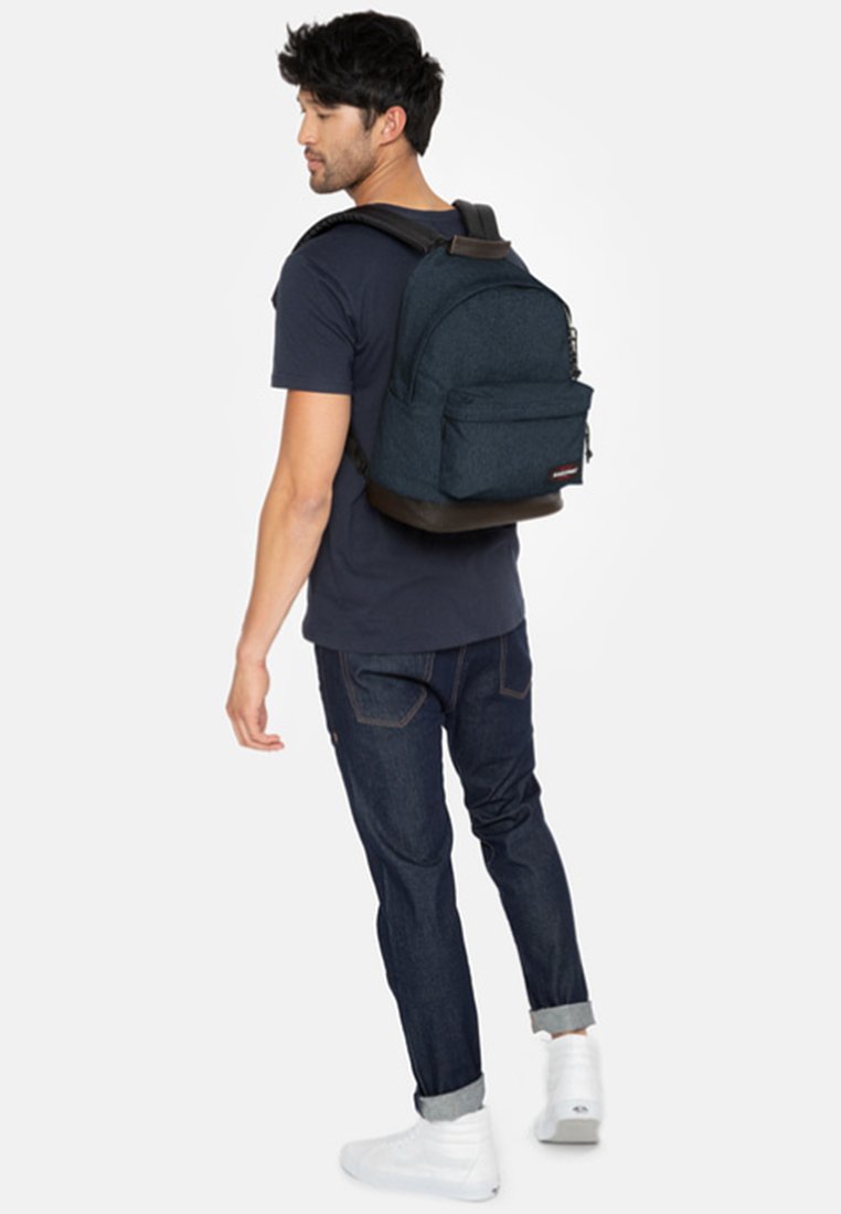 Mochila de tela azul con la parte superior redondeada, bolsillo frontal y base marrón; el modelo lleva una camiseta azul marino y jeans oscuros. Zapatillas blancas visibles.