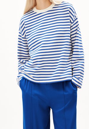 Persoon die een blauw-wit gestreept shirt met lange mouwen en een blauwe broek draagt, handen in de zakken, kijkend naar voren tegen een witte achtergrond.