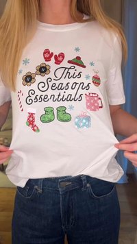 Maglietta bianca con grafiche festive che includono guanti, fiori, stivali e decorazioni natalizie in vari colori, con accenti scintillanti.