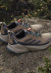Beżowe i granatowe buty outdoor z teksturowaną cholewką, zaprojektowane z myślą o stabilności, z elementami odblaskowymi oraz solidną podeszwą zapewniającą przyczepność.