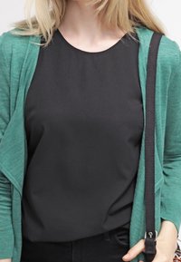 Vihreä neuletakki mustan topin päällä, jossa on sileä rakenne ja rento istuvuus. Mustat housut, joissa näkyy olkaintiedot.