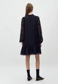 Rochie din dantela neagră cu o margine volanată, mâneci lungi transparente și guler înalt. Prezintă modele florale intricate și o potrivire lejeră.