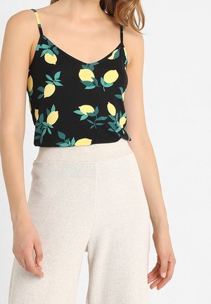 Kobieta w czarnej koszulce na ramiączkach z żółtym nadrukiem cytryn i białych, wysokich, teksturowanych spodniach na białym tle.