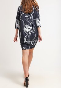 Vestido negro con patrones abstractos blancos, hecho de una tela suave. Presenta mangas tres cuartos y una silueta holgada. Tacones visibles.