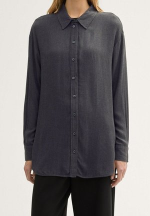 Chemise à manches longues gris foncé boutonnée portée par-dessus un pantalon noir sur une personne devant un fond clair uni.