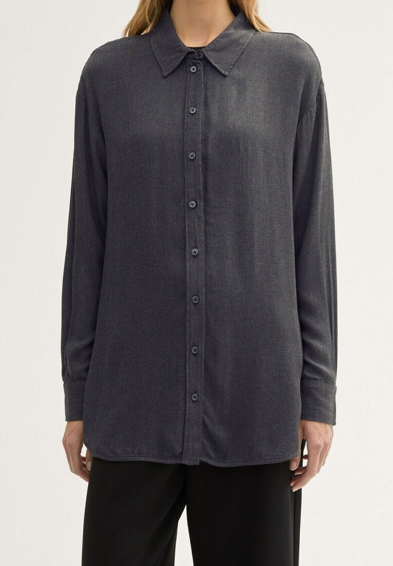 Chemise à manches longues gris foncé boutonnée portée par-dessus un pantalon noir sur une personne devant un fond clair uni.