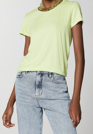 Osoba ubrana w jasną zieloną koszulę z krótkim rękawem, niebieskie jeansy z efektem acid wash oraz masywny przezroczysty zielony łańcuszkowy naszyjnik.