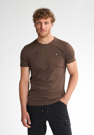 Man met krullend haar die een bruine korte mouwen T-shirt en een zwarte broek draagt, staand met één hand in de zak tegen een effen achtergrond.