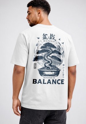 Man wearing white t-shirt with a dark blue bonsai tree graphic and text "Bonsai Rooted in Balance" on the back.