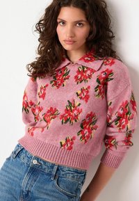 Roze gebreide trui met bloemenpatronen in rood en groen, met een kraag en ribgebreide manchetten. Gecombineerd met blauwe spijkerbroek.
