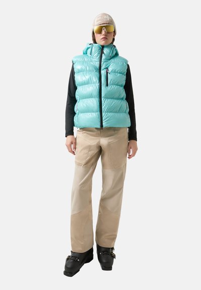 Puffweste in hellem Türkis mit Reißverschlusstaschen, kombiniert mit beigen Skihosen mit Paneeldetails und schwarzen Skischuhen. Abgerundet mit Sonnenbrille und Strickmütze.