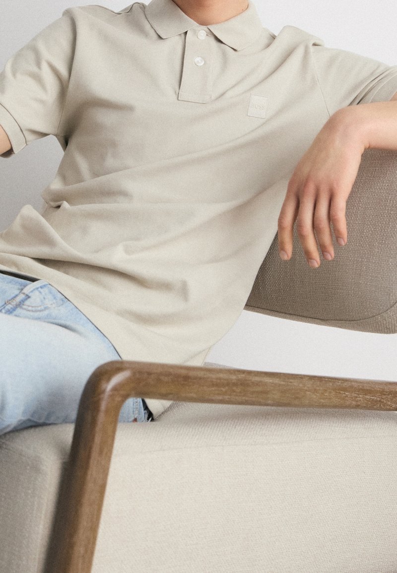 Polo en coton beige avec une patte de boutonnage à trois boutons et un écusson, associé à un jean bleu, assis sur un fauteuil texturé beige.