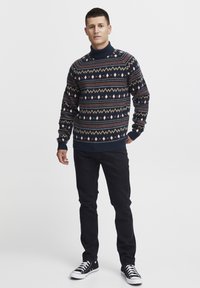 Pulover tricotat cu guler înalt, cu model în nuanțe de marină; are designuri geometrice în culori crem și rugină; purtat cu pantaloni negri și teniși.