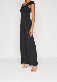 Schwarzer Jumpsuit aus weichem Material, mit einem verdrehten Oberteil, kurzen Ärmeln und weiten, fließenden Beinen. Getragen mit klaren, Riemchen-Absätzen.