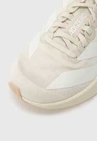 Heledat beežid spordijalatsid tekstuurse kangast pealse, valgete triipude ja kummisest tallaga. Omavad paeltega kinnitamist ja tikitud detaile.