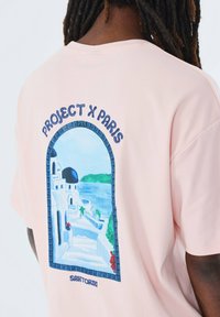 Camiseta de color rosa claro con un estampado gráfico de Santorini, arquitectura griega con techos en forma de cúpula, bordeada por un marco decorativo azul.