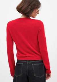 Punane kootud kampsun pika varrukaga, soonikute äärte ja hemiga, lihtsa disaini ja sileda tekstuuriga. Kandev tume denimist teksapükse.