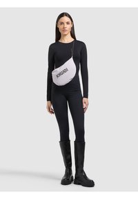 Femme portant un haut noir à manches longues, des leggings noirs, des bottes noires montantes jusqu'aux genoux et un sac bandoulière blanc avec le texte "KHUJO 2001".