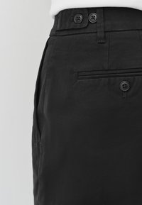 Pantalon noir avec un devant plat, deux boutons à la taille et une poche latérale. Le tissu semble lisse avec une légère brillance.