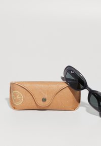 Juodos Ray-Ban katės akių formos saulės akinių pora gulinti šalia šviesiai rudos odinės Ray-Ban dėžutės su užsegimu, ant baltos paviršiaus.