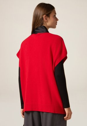 Rode gebreide trui met brede mouwen, geribbelde zoom en een gladde textuur. Draag je over een zwart longsleeve top, wat de gelaagde stijl benadrukt.