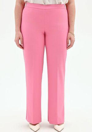 Femme portant un pantalon rose taille haute à coupe droite, associé à des chaussures blanches à bouts pointus et un chemisier blanc.